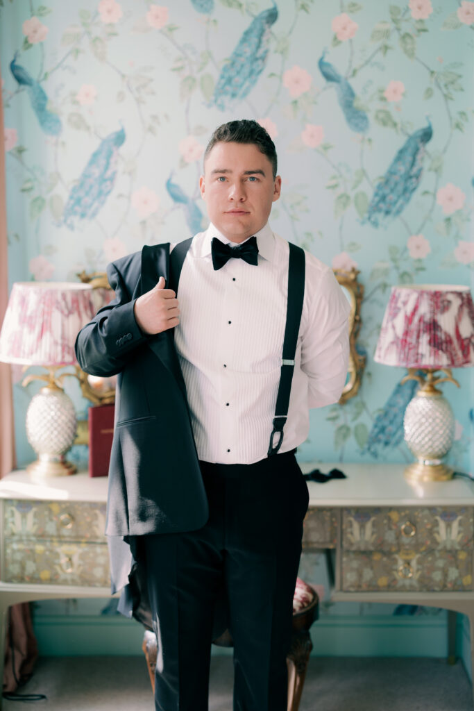 Groom in black suspenders and bow tie posing confidently against vibrant floral wallpaper backdrop