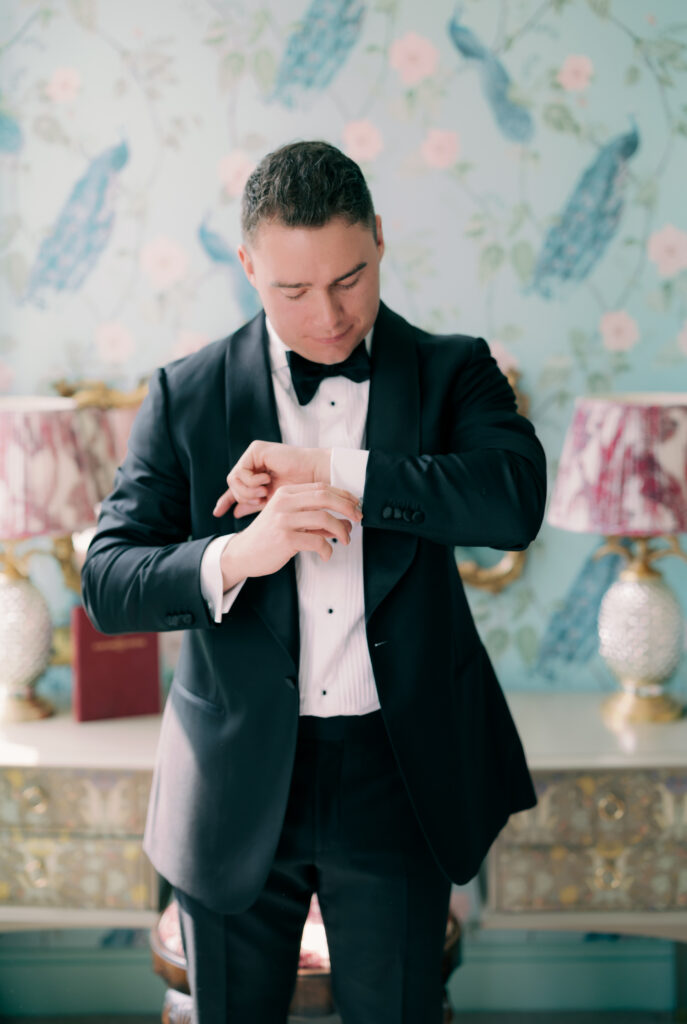 Groom in black suspenders and bow tie posing confidently against vibrant floral wallpaper backdrop